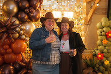 Load image into Gallery viewer, Two people in cowboy hats posing in front of a shimmer wall backdrop with balloons and flowers.