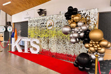 Load image into Gallery viewer, A shimmer wall with a white and gold color scheme, featuring large letters that spell 'KIS' in a decorative setting, with balloons and a red carpet.
