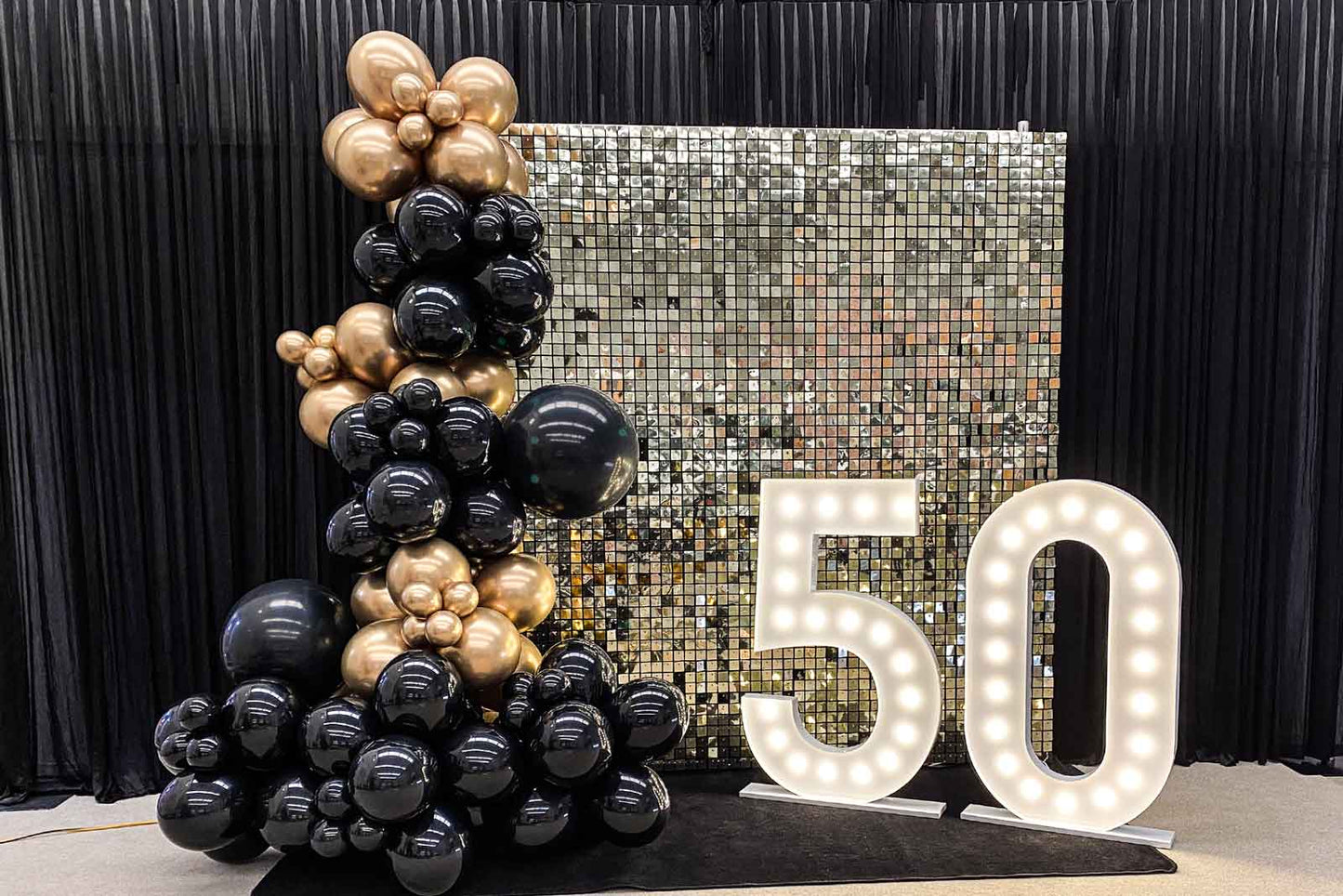 White gold shimmer wall with “50” marquee lights and black and gold balloon garland at an Auckland event