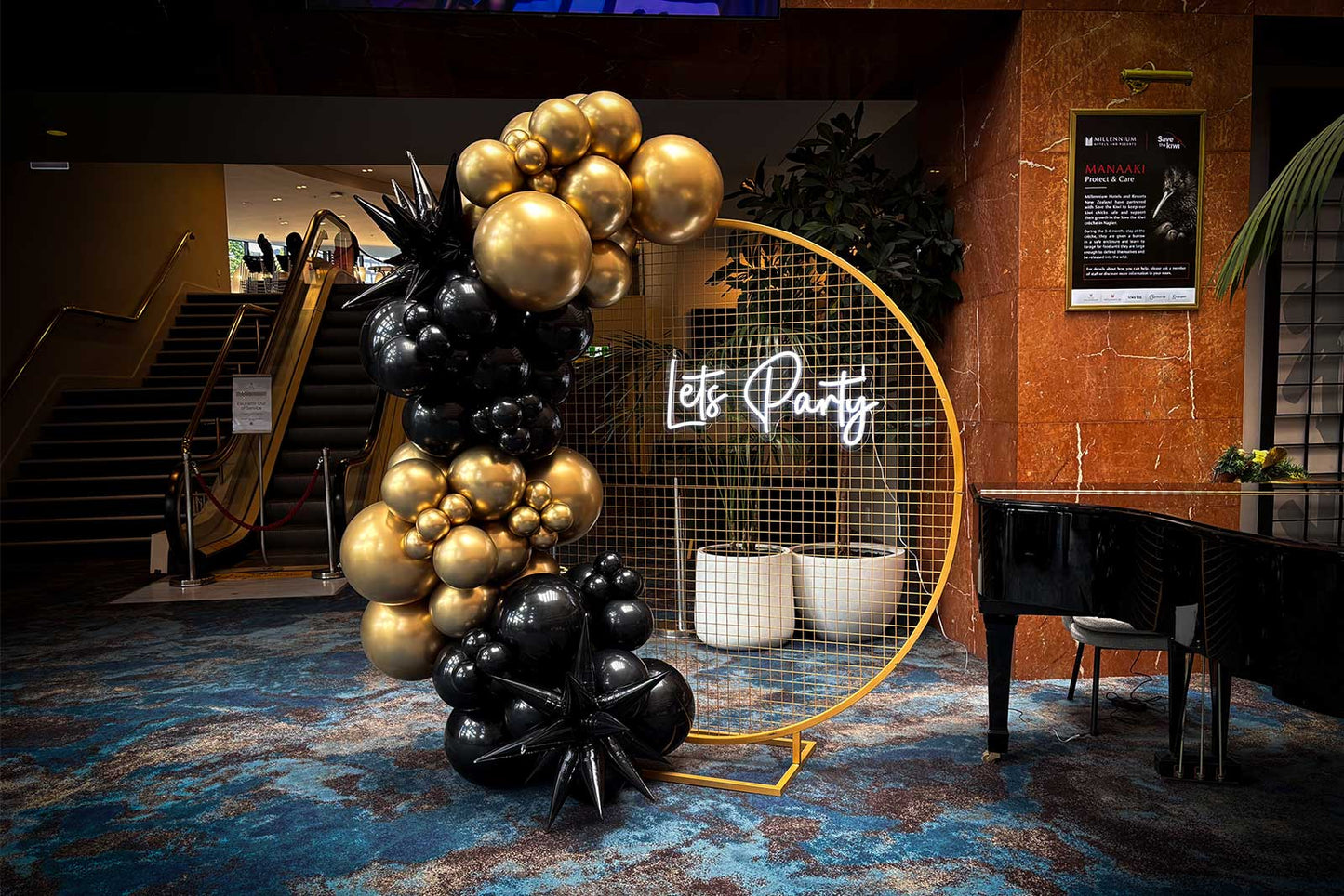 Black and gold balloon garland on a round gold mesh backdrop with a ‘Let’s Party’ neon sign, styled in a hotel lobby for an elegant event display.