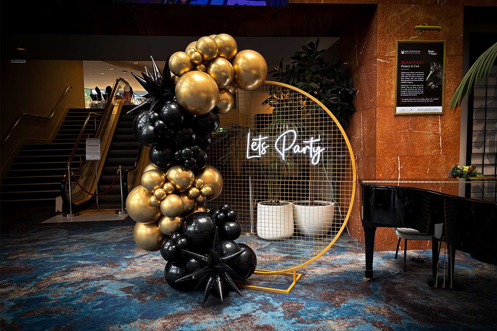Black and gold balloon garland on a round gold mesh backdrop with a ‘Let’s Party’ neon sign, styled in a hotel lobby for an elegant event display.