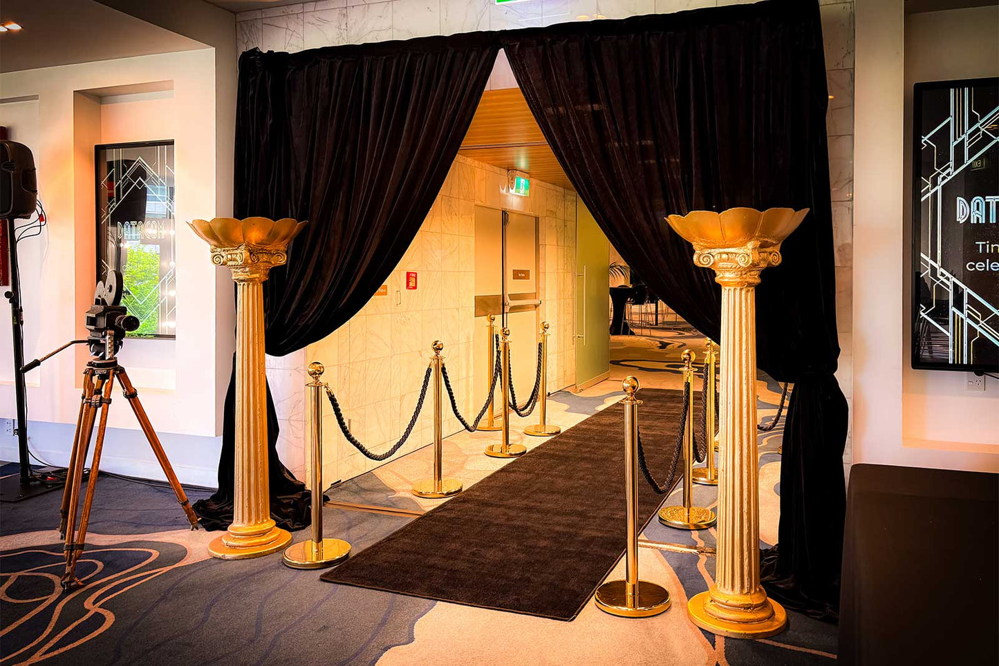 Elegant event entrance featuring black draping, a black carpet, walkway, gold columns and gold stanchions with black ropes, creating a luxurious Hollywood style arrival experience